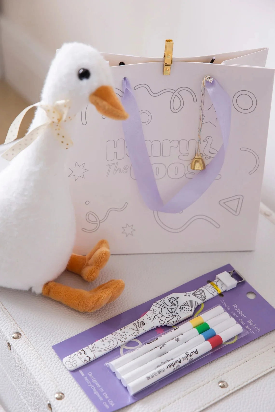 A plush Henry the Goose toy sits beside a branded gift bag with a lavender ribbon and a pack of non-toxic fabric markers for customizing a creative rubber watch.